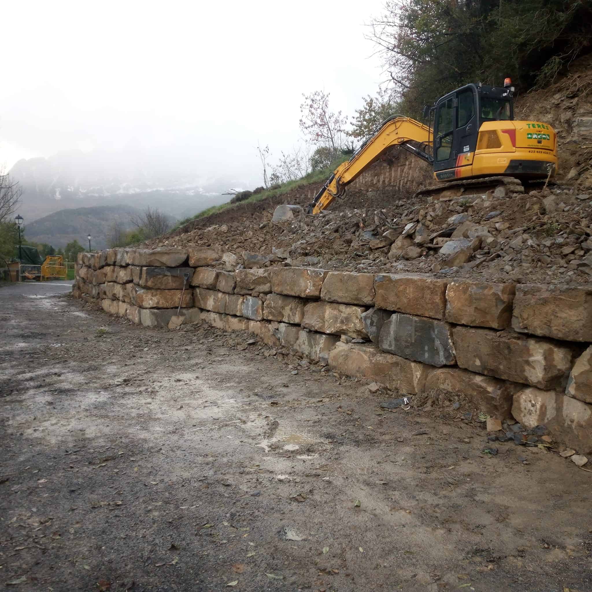 Retroexcavadora trabajando en construcción y reparación de muros de piedra para estabilización de terrenos en trabajos de obra civil y paisajismo.