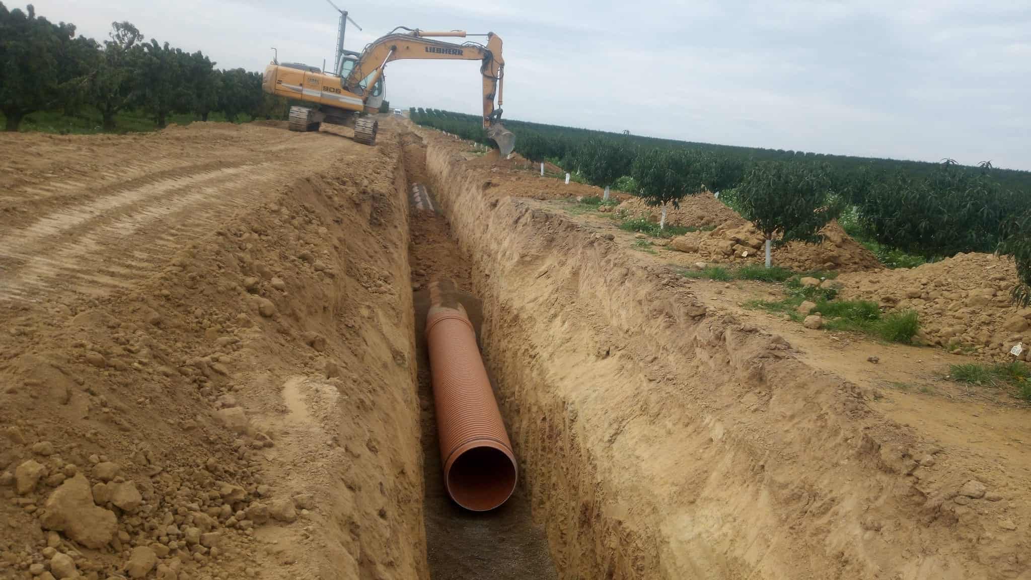 Tubería de alcantarillado instalada en una zanja profunda con maquinaria pesada, trabajando en trabajos y servicios de obras civiles y construcción en campos agrícolas.