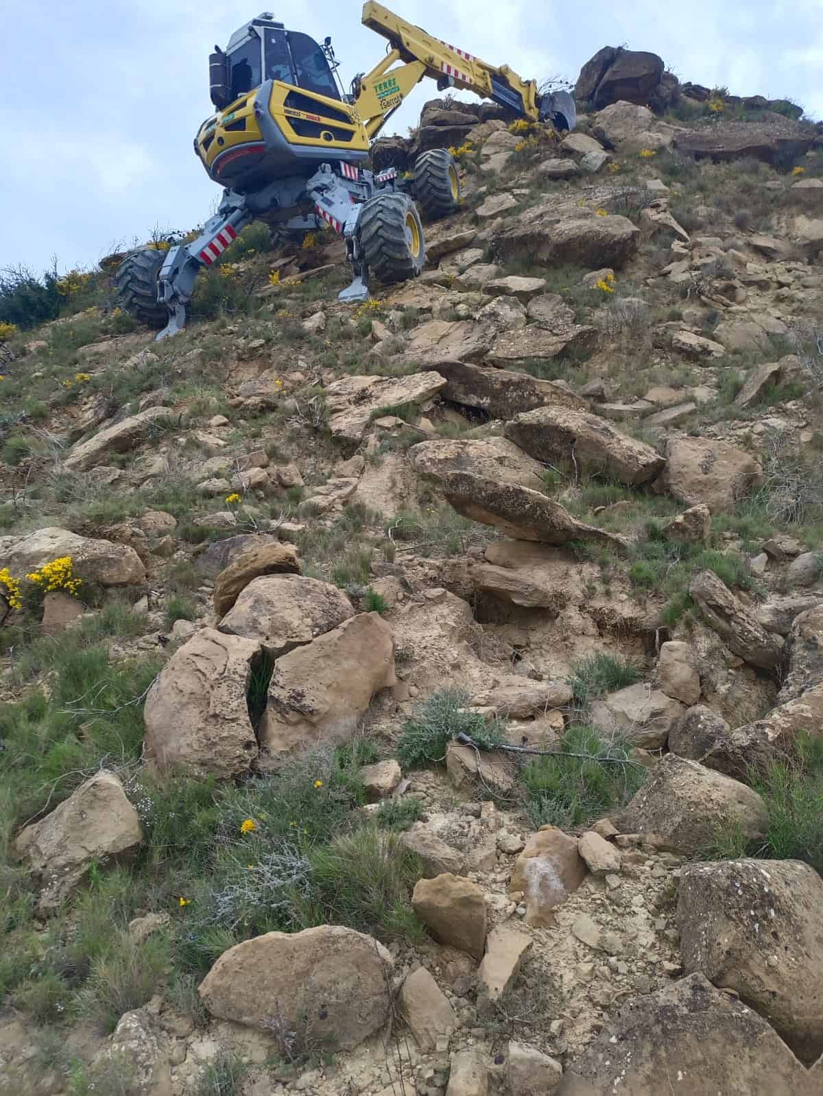 Maquinaria de excavación realizando trabajos en terreno rocoso y escarpado con piedras y vegetación, especializado en obras y servicios de terraceo y movimiento de tierras.