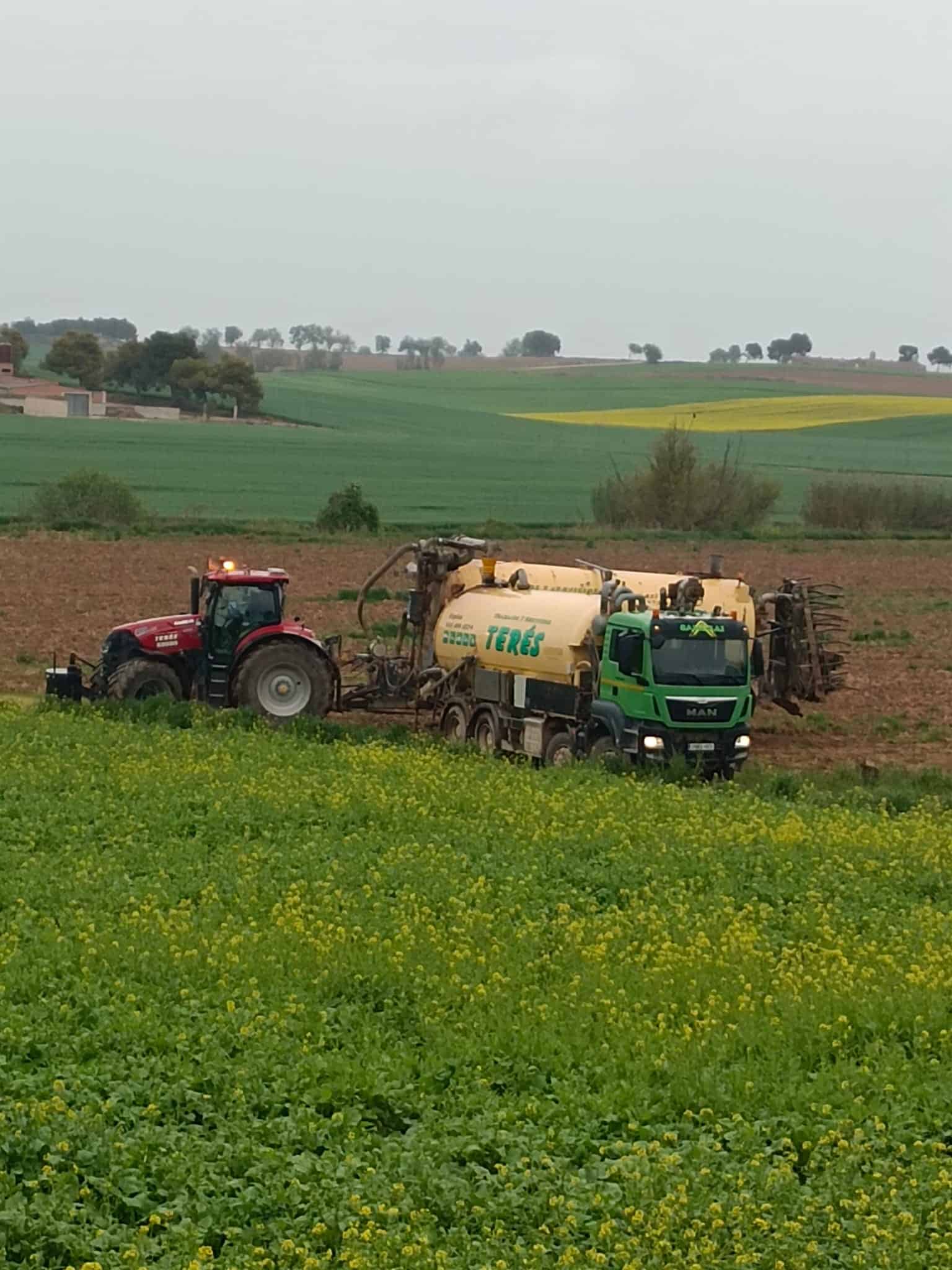 Maquinaria agrícola en campo de cultivo, tractor y camión de servicios agrícolas de Terés en movimiento en un paisaje rural de campos verdes y floridos.