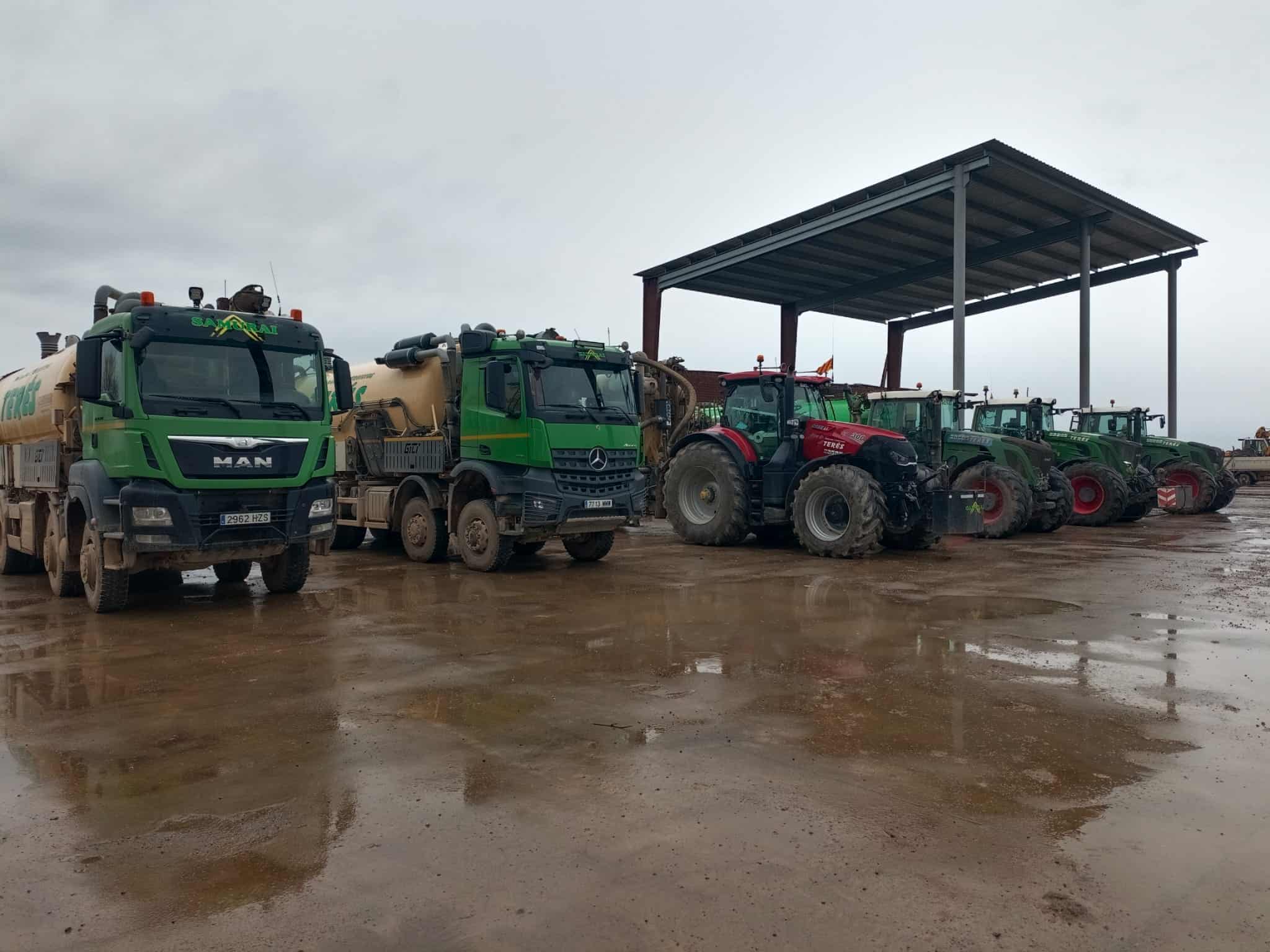Vehículos agrícolas y maquinaria de trabajos y servicios agrícolas en Terés, en un espacio al aire libre con suelo húmedo y un cobertizo metálico de fondo.